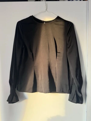 Svart blus med trumpetärm - Stilren svart blus med rund halsringning och långa trumpetärmar. Tillverkad i ett lätt, något glansigt tyg med struktur. Perfekt för dig som gillar en enkel men trendig look.
