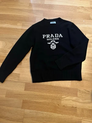 Svart stickad tröja från Prada - Snygg svart stickad tröja från Prada med vit logotyp och texten 'PRADA MILANO' framtill. Tröjan har rund halsringning, ribbade muddar och nederkant. Perfekt för dig som gillar exklusiva märken och klassisk stil.