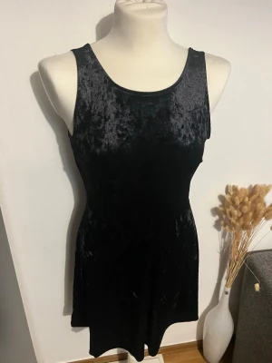 Svart sammetsklänning ärmlös - Snygg svart klänning i mjuk sammet med ärmlös design och rund halsringning. Klänningen har en figurnära passform upptill och lätt utställd kjol. Perfekt för dig som gillar en stilren och trendig look. Tyvärr storlekslappen är bortklippt 