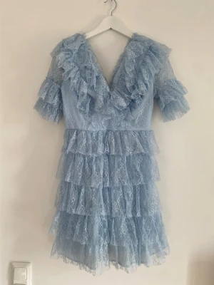 Ljusblå spetsklänning med volanger - Supersöt ljusblå klänning i spets med flera lager volanger och v-ringning. Klänningen har korta ärmar och romantiska detaljer med rysch längs både ärmar och urringning. Perfekt för dig som gillar en feminin och trendig look. klänningen har lite smink på 
