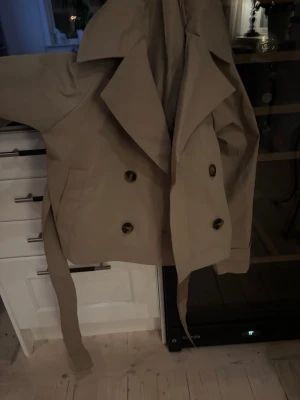 Kort beige trenchcoat med bälte - Snygg helt oanvänd trenchcoat i beige med dubbelknäppning och breda slag från Gina. Jackan har ett matchande bälte i midjan.