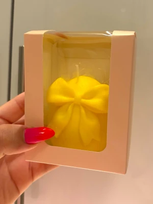 Gul dekorljus med rosett - Säljer ett gult dekorljus format som en rosett, levereras i en söt presentförpackning med genomskinligt fönster. Perfekt som inredningsdetalj eller present. Ljusets form är rund med en stor rosett framtill och det är tillverkat av vax.