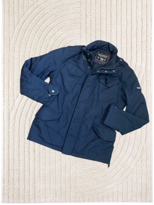 Woolrich field jacket  - Snygg marinblå jacka från Woolrich med flera praktiska fickor, hög krage och detaljer som knappar och dragkedja. Jackan har en klassisk design och är tillverkad i slitstarkt material, perfekt för dig som gillar stilrena ytterplagg.