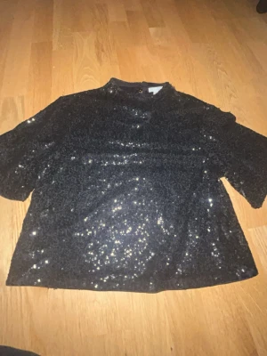 Svart paljettopp med kort ärm - Glittrig svart topp täckt med paljetter, har korta ärmar. Har nice material under paljetterna så att man inte känner dem. Två knäppen, är perfekt på en utekväll och lätt att matcha ihop me jeans och shorts. Har använt den två gånger men den har gått upp lite men inget håll i den och några paljetter har låsnat men inget man ser.