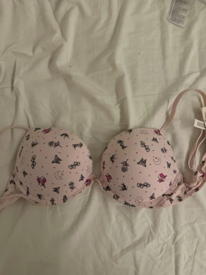 Ljusrosa bh med gulligt mönster - Söt ljusrosa bh med vadderade kupor och justerbara axelband. Bh:n har ett gulligt mönster med små figurer, månar och stjärnor i svart och rosa. Materialet känns mjukt och bekvämt, perfekt för dig som gillar lekfulla detaljer på underkläder.