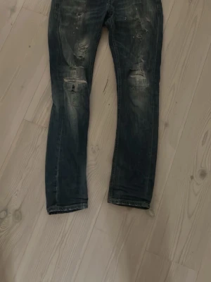 Mörkblå slitna jeans med detaljer - Säljer ett par mörkblå jeans med slitningar och distressed detaljer på både lår och knän. Jeansen har en rak passform och cool tvätt med ljusare partier. Perfekta för dig som gillar en avslappnad och trendig stil.