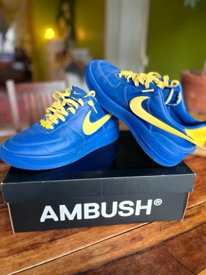 Nike x Ambush blå/gula air force  - Snygga sneakers i samarbete mellan Nike och Ambush. Skorna är blå med gula detaljer, inklusive gula skosnören och en gul swoosh. Tillverkade i läder med klassisk låg siluett och tydlig branding på plösen. Perfekta för dig som vill sticka ut med färg.