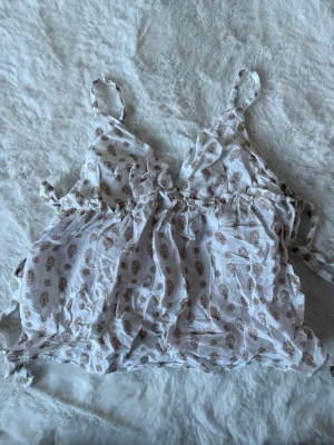 Linnetopp Nelly - Söt linnetopp i vitt med beige blommigt mönster och smala axelband. Toppen har volangdetaljer och knyt i sidorna. Storlek 32 från Nelly, endast testad.