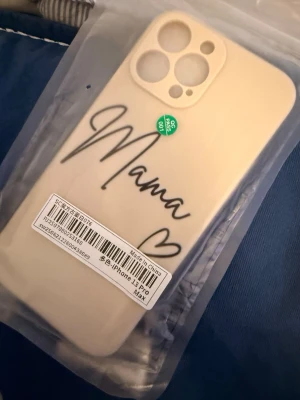 Beige mobilskal med text Mama - Snyggt beige mobilskal med svart text 'Mama' och ett hjärta, designat för iPhone 13 Pro Max. Skalets material är mjukt silikon som ger bra grepp och skydd. Perfekt för dig som vill ha ett personligt och stilrent skal till mobilen.