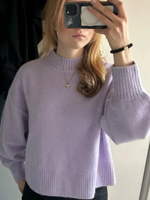 Ljuslila stickad tröja med ballongärm - Mysig ljuslila stickad tröja med ballongärmar och ribbade muddar vid ärmslut och nederkant. Tröjan har en högre rund halsringning och avslappnad passform. Perfekt för att addera färg till din garderob. Prislappen är kvar.