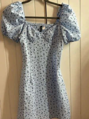 Ljusblå blommig klänning  - Söt ljusblå kortklänning från H&M med små mörkblå blommor och puffiga kortärmar. Klänningen har en utsvängd passform och resår vid axlarna för en bekväm look. Perfekt för varma dagar och ger en romantisk vibe.