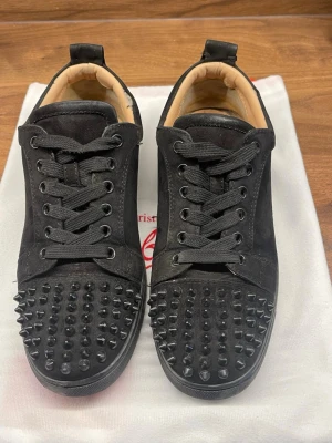 Svarta Louboutin sneakers med nitar - Svarta sneakers från Christian Louboutin i mocka med svarta nitar på tån och klassisk röd sula. Lågt skaft och svarta skosnören ger en clean och edgy vibe. Insidan är beige och skon har en unik design som sticker ut.