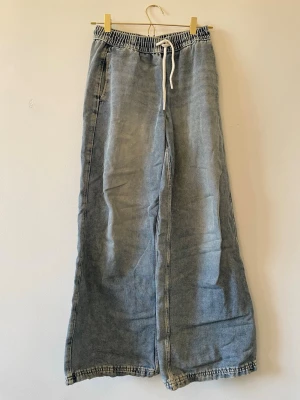 Vida ljusblå mjukisbyxor/jeans  med dragsko - Säljer ett par ljusblå mjukisbyxor/jeans med bred passform och resår i midjan samt vit dragsko. Byxorna har en avslappnad look, raka ben och en bakficka. Materialet ser ut som mjuk denim eller bomull, perfekta för chill dagar.