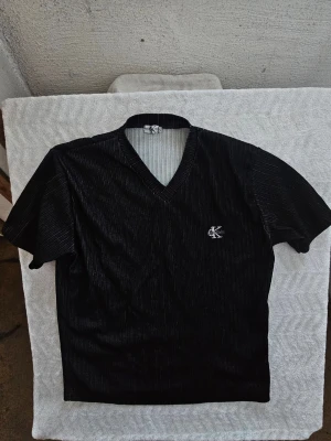 Svart v-ringad topp från Calvin Klein - Snygg svart topp från Calvin Klein Jeans med diskreta vita ränder och v-ringad hals. Kortärmad modell med broderad CK-logga på bröstet. Perfekt för en clean och stilren look.