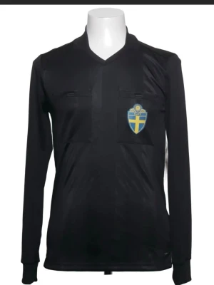 Svart långärmad domartröja SvFF - Svart långärmad domartröja från SvFF med två bröstfickor och det svenska fotbollsförbundets märke på vänster sida. Tröjan har V-ringning och är tillverkad i ett lätt, funktionellt material som passar perfekt till match eller träning.