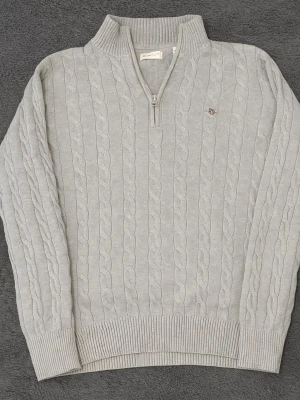 Ljusgrå kabelstickad tröja Gant - Kabelstickad ljusgrå tröja från Gant med halv dragkedja och ribbad krage. Tröjan har långa ärmar, ribbade muddar och en liten broderad logga på bröstet. Perfekt för dig som gillar klassisk och stilren look. Original pris på hemsidan 1700kr