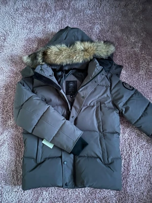 Canada goose jacka grå - Snygg grå dunjacka från Canada Goose med stor huva och pälskant. Jackan har raka, quiltade paneler, två sidofickor och en patch med logga på ärmen. Perfekt för kalla vinterdagar tack vare tjockt dunfoder och vindtätt material.