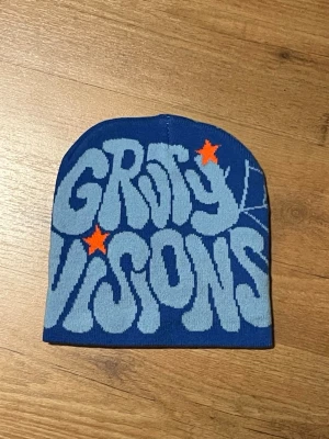 Blå mössa Groovy Visions - Snygg blå mössa med texten 'Groovy Visions' i ljusblått och två orange stjärnor. Mössan har en retroinspirerad design med rundad topp och är tillverkad i mjukt stickat material. Perfekt för dig som vill sticka ut med en cool accessoar.