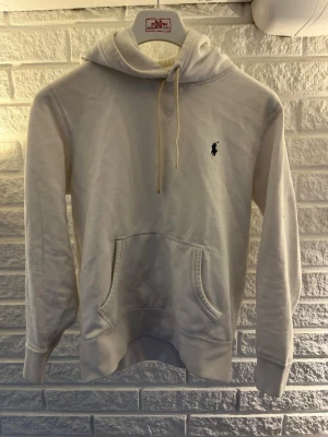 Vit hoodie från Polo Ralph Lauren - Säljer en klassisk vit hoodie från Polo Ralph Lauren med broderad, marinblå logga på bröstet. Hoodien har en stor och bekväm huva, känguruficka och ribbade muddar vid ärmslut och nederkant. Perfekt nu till sommaren för en lite mer uppklädd, men vardaglig stil.