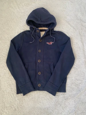 Mörkblå hoodie från Hollister - Mörkblå hoodie från Hollister med knappar framtill, dragsko i huvan och två fickor. Klassisk Hollister-logga broderad på bröstet. Tillverkad i mjukt bomullsmaterial som är skönt att ha på sig. Perfekt för en avslappnad och stilren look. Storleken är i Xl men passar mer som M/S. Tröjan har två laggade hål som då gör att priset blir billigare.