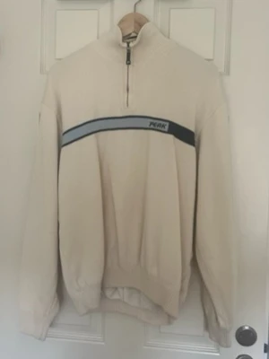 Beige halvzip tröja från Peak - Snygg beige stickad tröja från Peak med halv dragkedja och en bred mörkblå och ljusblå rand över bröstet. Tröjan har ribbade muddar och hög krage, perfekt för en chill och sportig look.