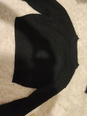 Svart stickad tröja med hålmönster - Säljer en svart stickad tröja med trendigt hålmönster och ribbade muddar. Tröjan har lång ärm och en croppad passform som passar perfekt till höga jeans eller kjol. Materialet känns mjukt och följsamt, perfekt för lager på lager.