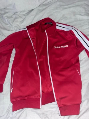 Röd tracksuit från Palm Angels - Snygg röd track jacket från Palm Angels med vita detaljer och dragkedja framtill. Jackan har vita ränder längs ärmarna och logga på bröstet. Tillverkad i polyester och bomull för en sportig och bekväm känsla. Perfekt till streetwear eller träning.