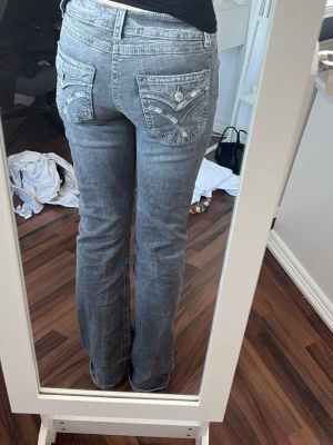 Grå bootcut jeans med detaljer - Säljer ett par grå bootcut jeans med snygga broderade bakfickor och dekorativa knappar. Jeansen har låg midja och klassisk femficksmodell. Perfekta för dig som gillar en avslappnad men ändå trendig stil. (Pris går att diskutera)