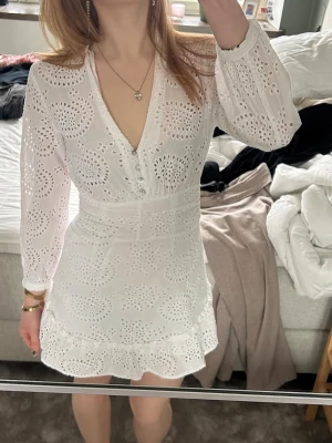 Studentklänning, vit kort klännning med mönster - Supersöt vit kort klänning med broderie anglaise, v-ringning och knappar framtill. Klänningen har långa ärmar, markerad midja och volangkant nertill. Perfekt för student eller konfirmation 💖