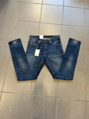 Blå slim fit jeans från tiger of sweden W29 L34 - Snygga blå slim fit jeans från tiger of sweden med klassisk femficksdesign och lätt slitna detaljer på benen. Jeansen har normal midja, dragkedjegylf och är tillverkade i ett stretchigt denimtyg för skön passform. Perfekta för en trendig look.