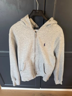 Grå zip hoodie från Polo Ralph Lauren - Snygg grå hoodie med dragkedja från Polo Ralph Lauren. Klassisk design med huva, fickor framtill och den ikoniska lilla loggan broderad på bröstet. Perfekt till jeans eller joggers. Tillverkad i mjukt bomullsmaterial. 
