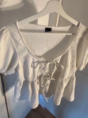 Vit blus med knyt och puffärm - Supersöt vit blus från Gina Tricot med puffärmar och två knyt framtill. Blusen har en lätt rynkad struktur och volang nedtill som ger en romantisk vibe. Perfekt till jeans eller kjol för en trendig look.