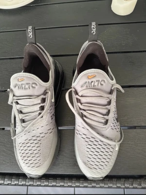 Nike Air Max 270 grå sneakers - Nike Air Max 270 sneakers i grå mesh med svarta och orange detaljer. Skorna har en stor svart Air Max-häl, vita och orange swoosh-loggor på sidorna och AIR270-tryck på plösen. Lågt skaft och ventilerande material gör dem perfekta för en sportig look.