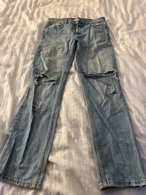 Ljusa slitna jeans midwaist från Zara - Säljer ett par ljusblå jeans från Zara med raka ben och slitningar över båda knäna. Jeansen har klassisk femficksdesign och är tillverkade i denim. Perfekta för en avslappnad och trendig look.