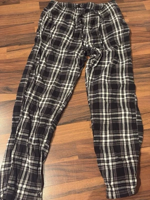Rutiga pyjamasbyxor i svartvitt - Säljer ett par sköna pyjamasbyxor med svartvitt rutmönster. Byxorna har resår i midjan med dragsko för bästa passform och är tillverkade i mjukt tyg, perfekt för chillkvällar hemma. Klassisk och avslappnad modell som passar alla som gillar mysiga hemmakläder. Lite korta efter att ha tvättats. (Pris går att diskutera) 