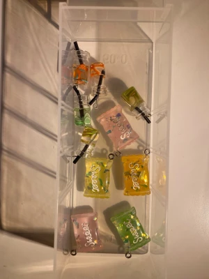 Färgglada godis- och dryckesberlocker - Säljer ett gäng söta berlocker i form av godispåsar och små dryckesflaskor i plast. Färgerna är blandade: rosa, grön, gul och orange. Perfekta att använda till smyckestillverkning, t.ex. som hängen till halsband, armband eller örhängen.