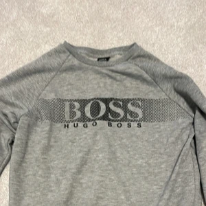 Grå sweatshirt från Hugo Boss - Snygg grå sweatshirt från Hugo Boss med stort svart logotryck över bröstet. Tröjan har rund halsringning och långa ärmar. Perfekt för en stilren och avslappnad look.