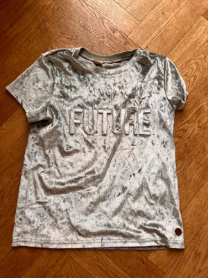 Silvrig sammetst-shirt med 3Dtext - Säljer en silvrig t-shirt i mjuk sammet med texten FUTURE i relief på bröstet. T-shirten har korta ärmar och rund hals. Perfekt för dig som vill sticka ut med en unik och trendig look. T-shirten är egentligen för barn ålder 10 men kan passa XS också! Skriv gärna för mått! 💕