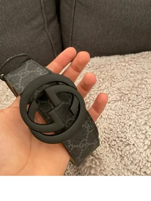 Gg bälte - Snyggt svart Gg bälte med stor matt svart dubbel-G logga som spänne. Bältet har ett grått monogrammönster över hela remmen och insidan är beige. Perfekt accessoar för att lyfta vilken outfit som helst. Kommer utan justerings bandet