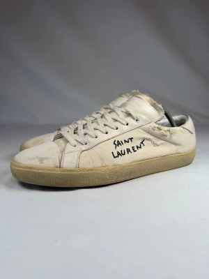 Saint Laurent vita sneakers med text - Vita sneakers från Saint Laurent med handskriven svart text på sidan. Skorna har lågt skaft, beige sula och slitna detaljer för en vintage look. Tillverkade i läder med klassisk snörning och rund tå.