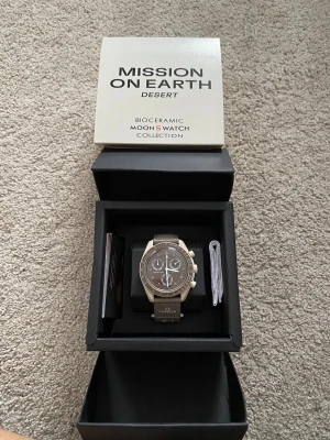 Omega x Swatch MoonSwatch Desert - Säljer en Omega x Swatch MoonSwatch Mission on Earth Desert från Bioceramic MoonSwatch Collection. Klockan har en beige och brun färgskala, tachymeter och flera subdials. Armbandet är matchande beige med Omega-logga. Perfekt för dig som gillar unik design.