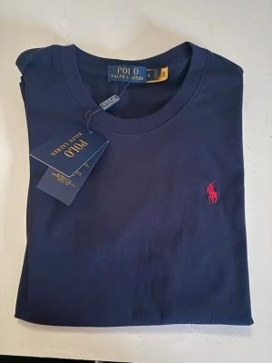 Marinblå t-shirt från Polo Ralph Lauren - Snygg marinblå t-shirt från Polo Ralph Lauren med klassisk rund hals och ikonisk röd logga broderad på bröstet. Tillverkad i mjuk bomull för skön känsla och stilren look. Perfekt till jeans eller shorts för en clean outfit. Rekommenderar starkt perfekt nu till sommaren kommer med allt