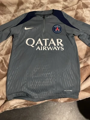 Grå PSG träningströja från Nike - Snygg grå träningströja från Nike med PSG-logga och Qatar Airways-tryck på bröstet. Tröjan har halv dragkedja, lång ärm och är tillverkad i Dri-FIT-material som andas. Perfekt för fotbollsträning eller gymmet.