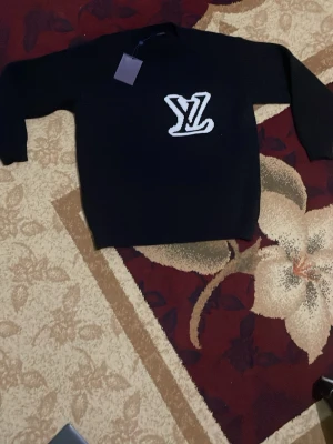 Svart tröja med LV-logga - Snygg svart tröja från Louis Vuitton med stor vit LV-logga på bröstet. Tröjan har rund halsringning och långa ärmar. Perfekt för dig som gillar streetwear och vill ha en statement-piece i garderoben.