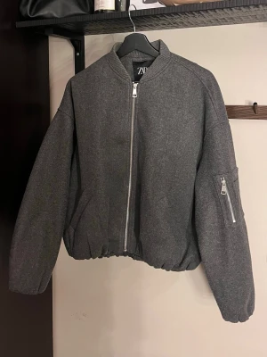 Grå bomberjacka från ZARA - Snygg grå bomberjacka från ZARA med silverfärgad dragkedja framtill och på ärmen. Jackan har en klassisk bombermodell med ribbad krage och elastisk nederkant. Tillverkad i ett mjukt tyg som ger en clean och modern look.