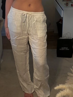 Vita linnebyxor med dragsko - Säljer ett par vita linnebyxor med avslappnad passform och dragsko i midjan. Byxorna har raka ben och elastisk midja för extra komfort. Perfekta för en chill och stilren look. Materialet är luftigt linne som känns svalt och skönt.