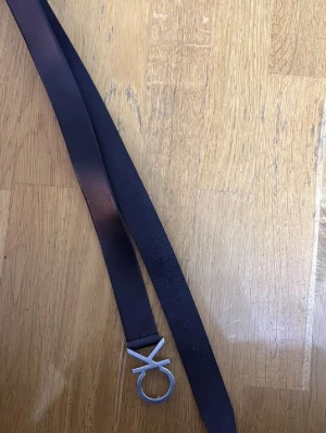 Svart bälte Calvin Klein - Snyggt svart bälte från United Colors of Benetton med ett unikt silverfärgat spänne i form av deras ikoniska logga. Bältet är smalt och tillverkat i skinn, perfekt för att lyfta vilken outfit som helst.