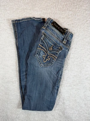 Rock Revival bootcut jeans storlek 27 - Snygga blå jeans från Rock Revival med dekorativa sömmar och broderad bakficka. Jeansen har klassisk femficksdesign, raka ben och slitningar som ger en cool look. Materialet är denim med detaljer i kontrastfärg. Perfekt för dig som gillar streetstyle.