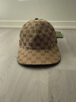  Beige keps med GG-mönster - Säljer en beige keps med det klassiska GG-monogrammet över hela kepsen. På sidan finns den ikoniska gröna och röda randen. Kepsen har böjd skärm och är tillverkad i canvasmaterial. Perfekt för dig som vill ha en lyxig och trendig accessoar.