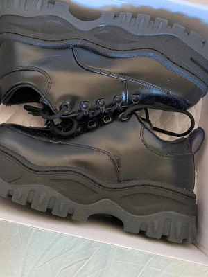 Eytys angels sneaker  - Säljer ett par chunky svarta kängor med kraftig, mönstrad sula och snörning. Skorna har en blank finish och robust design som ger en cool och edgy look. Perfekta för dig som gillar streetstyle och vill sticka ut. Materialet är troligtvis konstläder.